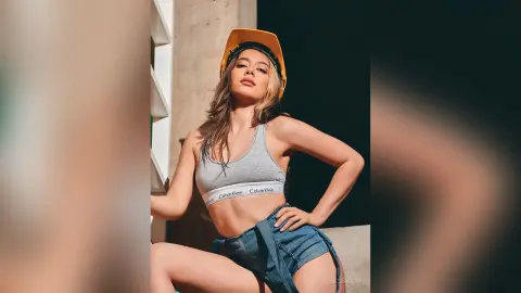 JessieLiu poses in a grey sports bra and denim shorts while wearing a yellow cap.