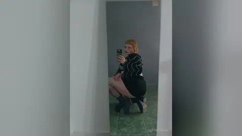 Redheaded woman in a black striped sweater taking a moody mirror selfie in a minimalist room.