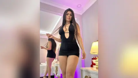 Young woman in a sleek black mini dress posing confidently in a brightly lit bedroom.