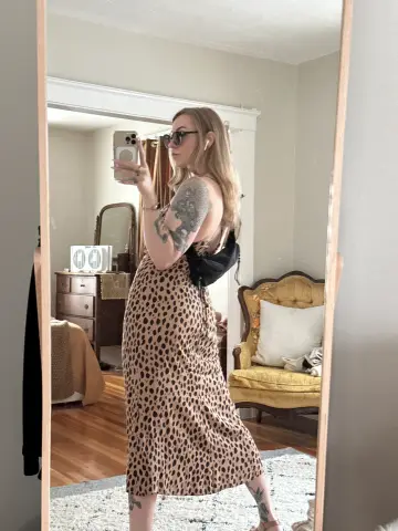 Woman in leopard print midi dress taking a mirror selfie in a cozy, sunlit bedroom.