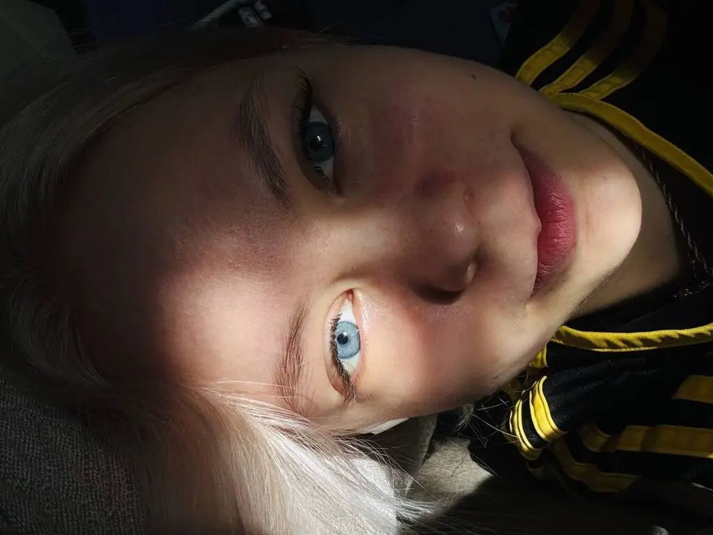 Close-up portrait of a blonde woman with blue eyes wearing a black and yellow striped shirt in soft lighting.