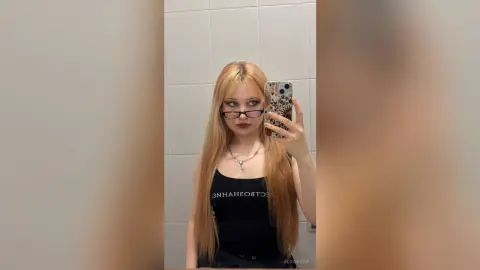 EloiseGow is a blonde woman with glasses wearing a black tank top, taking a mirror selfie in a tiled bathroom.