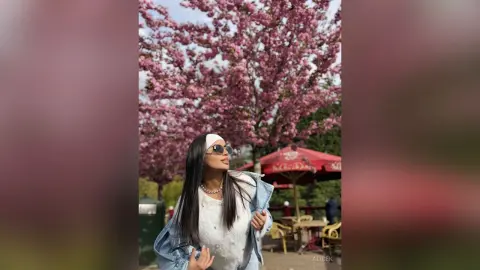Stylish woman in white top and denim jacket smiling under blooming pink cherry blossom trees in a sunny park.
