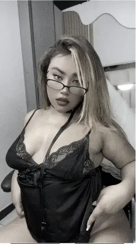 Curvy woman in black lace lingerie and glasses posing confidently in a bedroom setting.