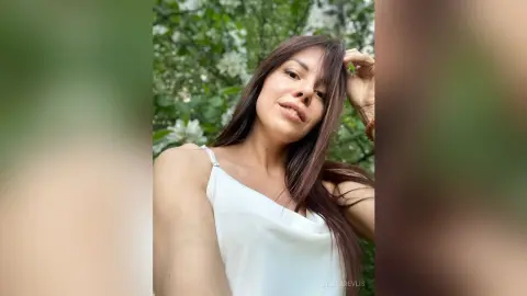Smiling woman in a white sleeveless top posing in a lush green garden setting.