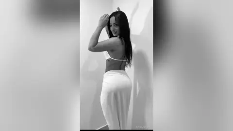 Elegant woman in a white bikini and flowing skirt posing against a minimalist wall in a moody black and white portrait.