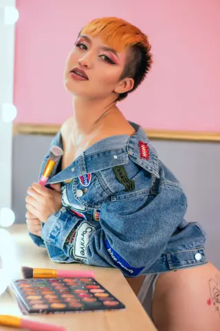 Stylish woman with short blonde hair in a denim jacket posing by a makeup palette in a pink room.