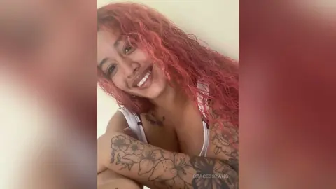 Smiling woman with wavy pink hair and floral arm tattoos wearing a white tank top in a bright indoor setting.