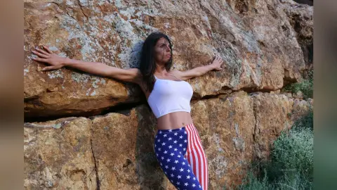 Woman in patriotic star and stripe leggings leaning against a rugged rock wall in a peaceful outdoor setting.