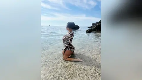 Woman in a zebra print swimsuit kneeling on a sunny beach by the ocean waves.