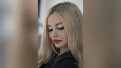 Blonde woman with silver eye makeup wearing a black shirt in a soft, moody portrait setting.