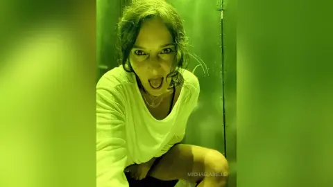 MichaelaBelle leans forward with her mouth open, wearing a white t-shirt and looking directly at the camera.