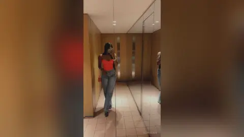 Young woman in a red tank top and blue jeans taking a mirror selfie in a modern elevator hallway.