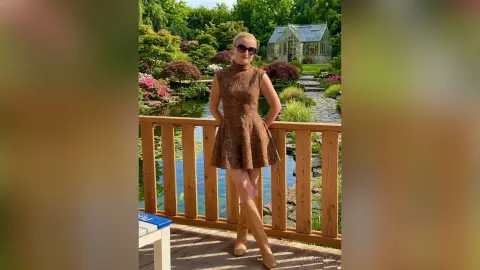 Blonde woman in a brown sleeveless mini dress standing on a wooden deck by a garden pond.