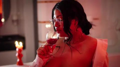 Young woman in a white shirt sipping red wine in a moody, dimly lit room with warm candlelight.