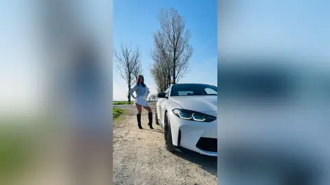 DyandraEve stands outdoors next to a white car, wearing a light blue long-sleeve top, a short white skirt, and black knee-high boots.