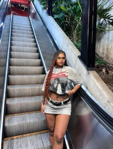 Stylish woman in a graphic t-shirt and denim mini skirt posing on an outdoor escalator near lush greenery.