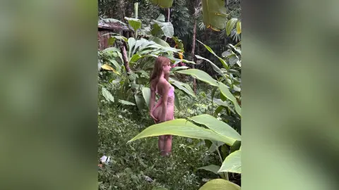 Young woman in a pink bikini standing in a lush tropical jungle garden with large green banana leaves.