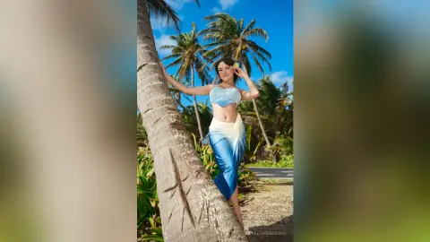 Young woman in a blue bikini top and white sarong leaning against a palm tree on a sunny tropical beach.
