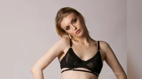 Blonde woman in a black lace bra posing against a neutral studio background with a confident mood.