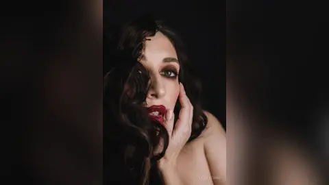 Glamorous woman with wavy dark hair and bold red lipstick posing in a moody, dark studio setting.