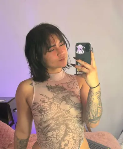 Young woman with dark hair and tattoos wearing a sheer dragon print bodysuit in a soft purple lit room.