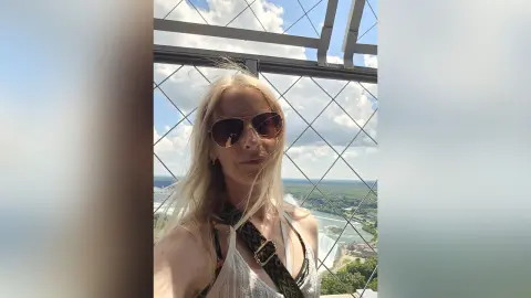 Blonde woman in sunglasses enjoying a sunny view from a high balcony overlooking a scenic landscape.
