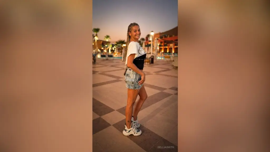 DelliaSmith โ DelliaSmith smiles while standing outdoors in a white t-shirt, denim shorts, and white sneakers on a tiled walkway.