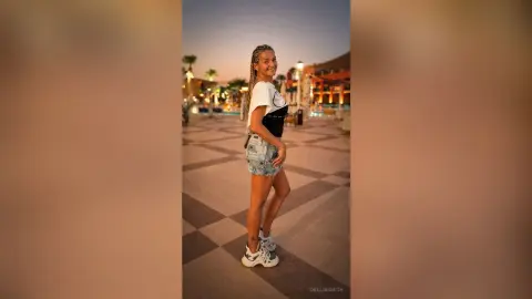 DelliaSmith smiles while standing outdoors in a white t-shirt, denim shorts, and white sneakers on a tiled walkway.