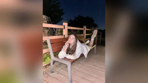 Cheerful young woman in a white sweatshirt relaxing on a wooden park bench at night.