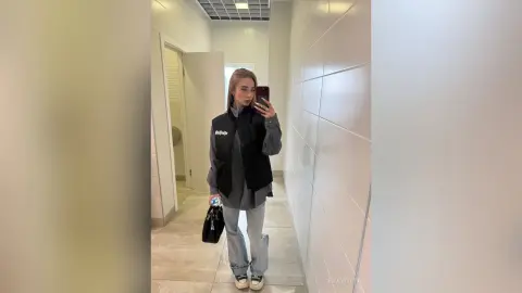 Young woman in a black vest and baggy jeans taking a mirror selfie in a modern tiled hallway.