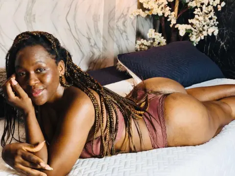 Smiling Black woman with long braids in mauve lingerie relaxing on a white bed in a bright bedroom.