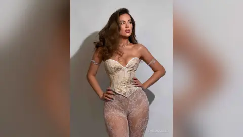 EvelynMills poses hands on hips wearing a cream corset and sheer lace tights against a plain background.