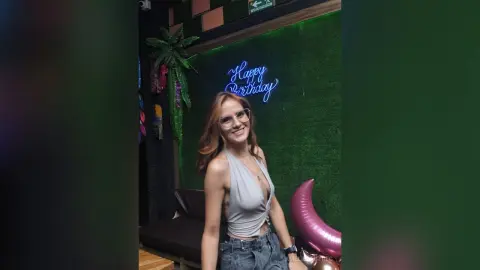 AmelieLautrec smiles wearing glasses and a low-cut grey tank top, posing in front of a green grass wall with a Happy Birthday sign.