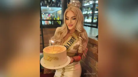 JessycaPage is a blonde woman wearing a crown and a birthday sash while holding a lit birthday cake.