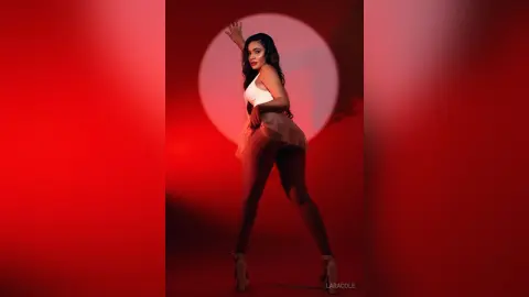LaraCole poses in a white tank top and light pink skirt against a bright red circular light, showing off her long dark hair and legs.
