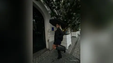 Stylish woman in a black outfit holding a black umbrella on a cobblestone street near white planters.