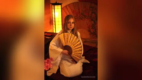 Young woman in a white kimono holding a traditional folding fan in a warm, dimly lit Japanese-style room.
