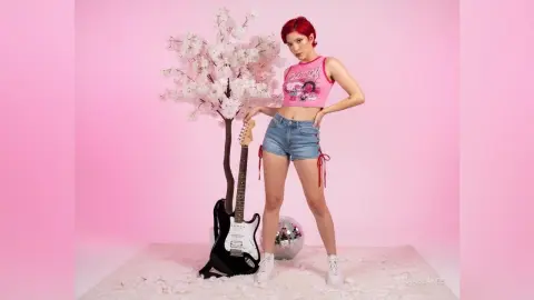 Red-haired woman in a pink graphic tank top and denim shorts posing by a cherry blossom tree against a pink backdrop.