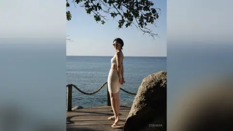 Stylish woman in a white bikini and skirt posing on a wooden pier by the ocean under a clear blue sky.
