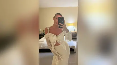 EmmaFitzgerald poses in a mirror wearing a white lace outfit with cutouts, showing her midriff in a bedroom setting.