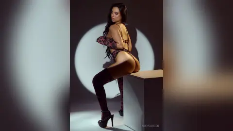 SofyLauren poses in a red patterned lingerie set and thigh-high stockings, showing her bare back and buttocks against a dark studio background.