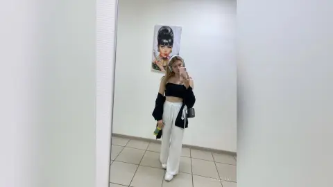 Young woman in a black crop top and white trousers taking a mirror selfie in a bright, modern room.