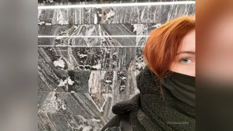Redheaded woman in a black fleece jacket and face mask standing against a textured marble wall.