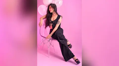 SofiCarvalo sits on a clear chair wearing black sleeveless clothes against a pink background.