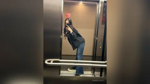Young woman in a black long-sleeve shirt and blue jeans taking a mirror selfie in a modern elevator.