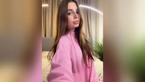 GertudeCova poses in a pink sweater with her long brown hair down in a bedroom setting.