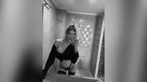 AriaVex poses in a black off-the-shoulder top and black thong in front of a tiled bathroom mirror.