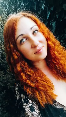 Jill_Stevens smiles with her long, wavy red hair against a dark green leafy background, wearing a black lace top.