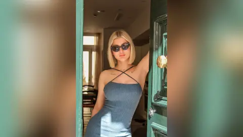 DaisyLin poses in a tight blue dress and sunglasses while standing in a doorway.